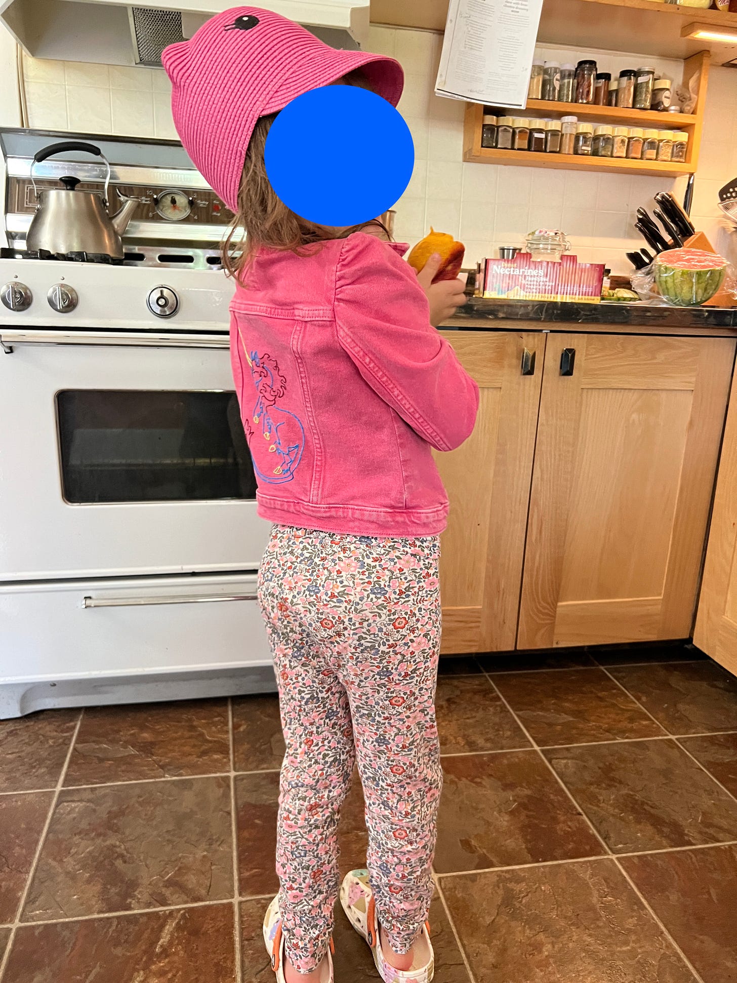 Photo of a five-year-old wearing a pink denim jacket with a unicorn embroidered on the hat. They are also wearing a pink 'cat' hat with little ears and cartoon eyes and flowery pink PJs