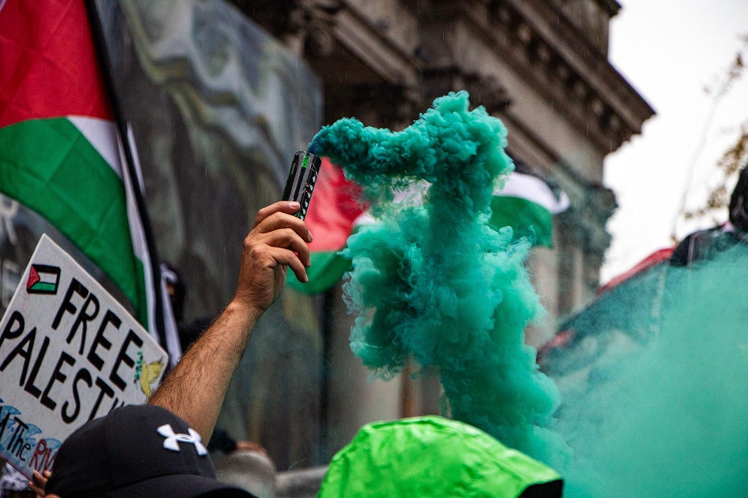 A pro-Palestine protester holds up a green smoke canister with a placard behind them reading "Free Palestine"
