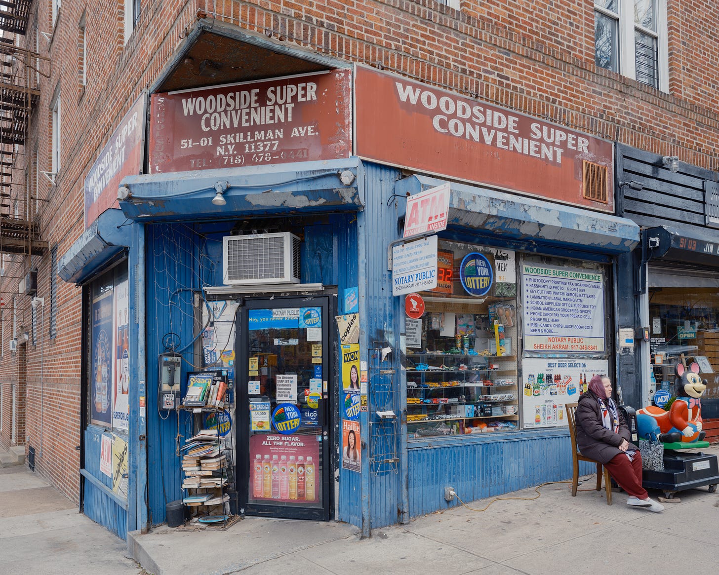 An older woman with a headscarf sits outside Woodside Super Convent store nest to a ride on mouse machine