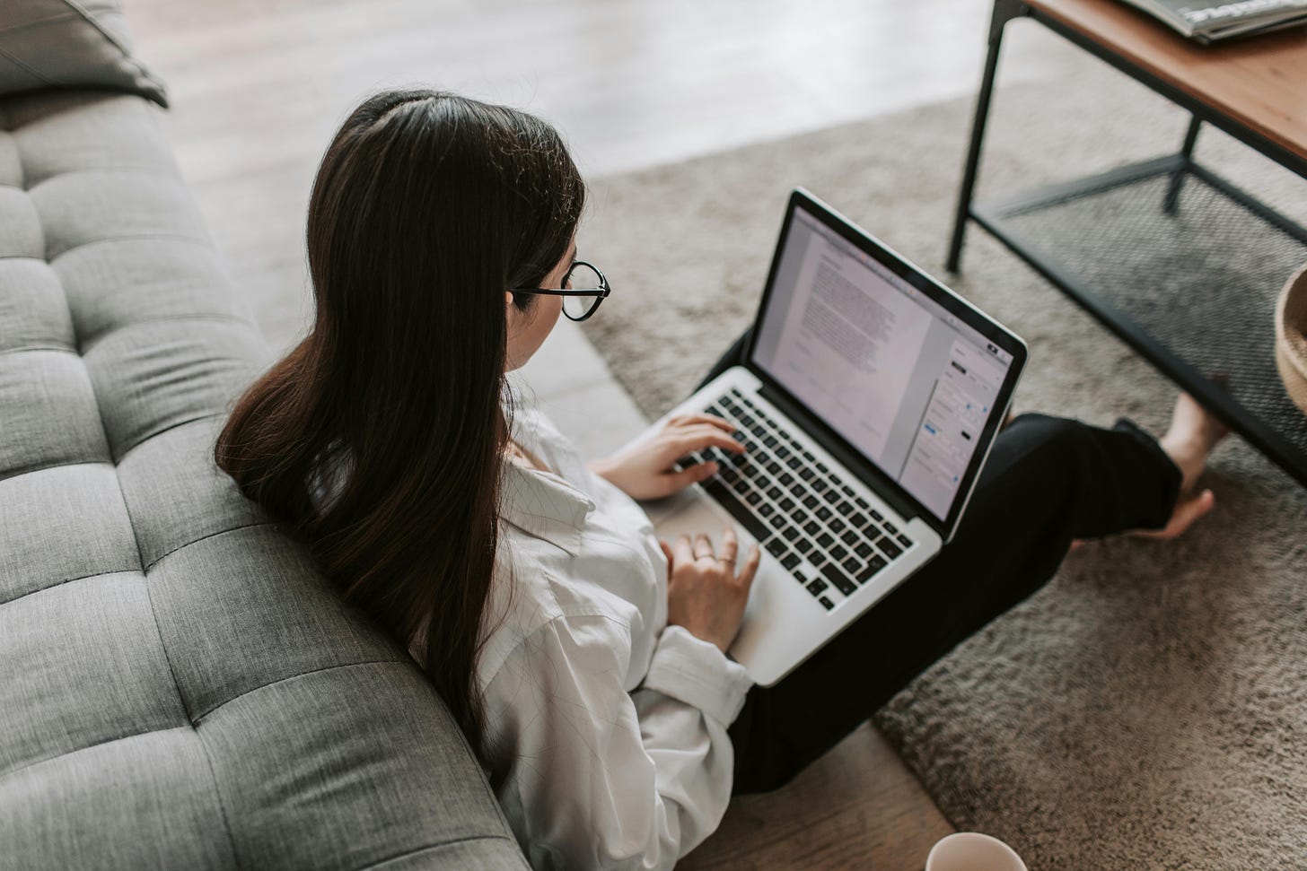 A woman sitting on the floor next to a gray sofa, working on a laptop. She has long brown hair and is wearing glasses and a white shirt. She appears focused, typing on the keyboard with both hands. The laptop screen displays a document editing software. A coffee cup is placed on the floor nearby, and a wooden table is partially visible in the background.