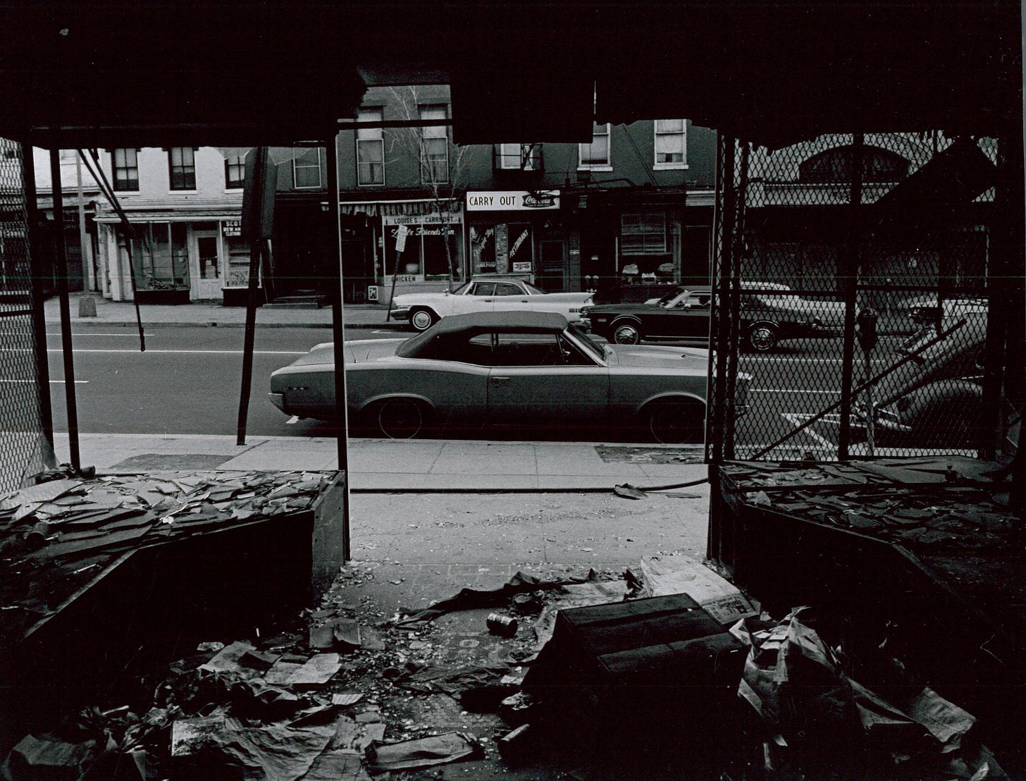 Photo taken from the inside of a store that appears to have been looted and largely destroyed. Businesses across the street appear similarly destroyed. Luxury cars are on the street, providing significant situational contrast.