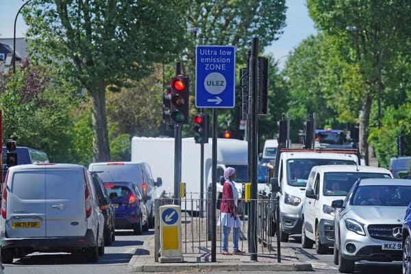 A half-dozen cars and vans are stopped at a red light beside a blue street sign that reads "Ultra low emission zone."