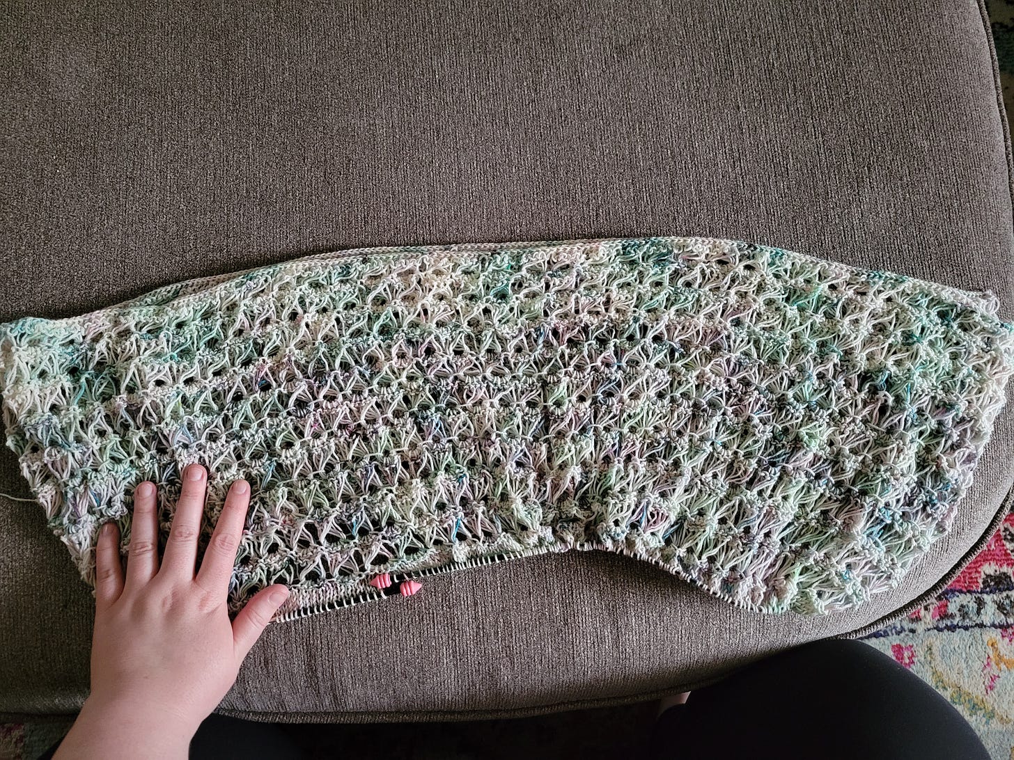 A panel of knitted broomstick lace in a watercolor-style variegated yarn in greens, blues, purples, and white. The panel is spread out on a gray ottoman and a white woman's hand is on top of it.