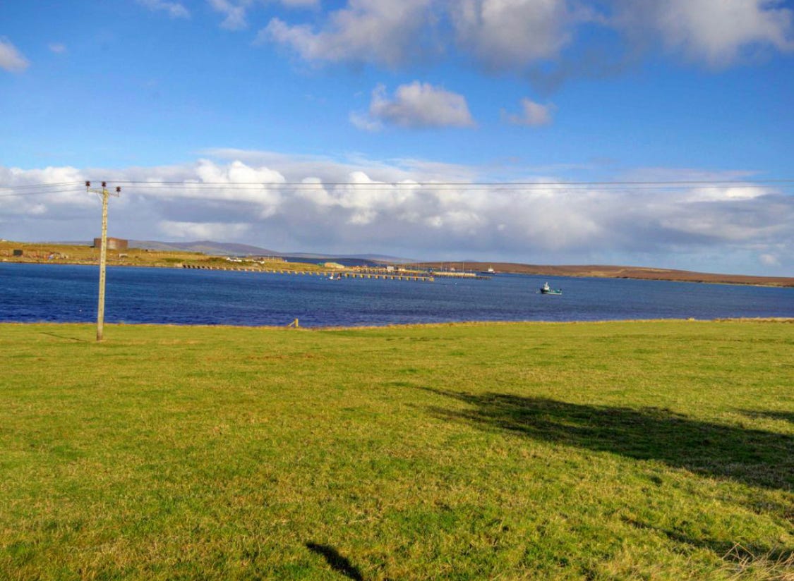 Landscape image of a bay, in the forgeound is a green field, in the background is the land on the other side of the bay. 