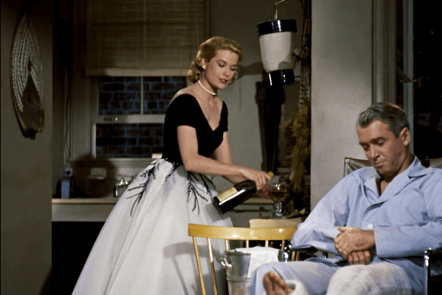 Grace Kelly as Lisa Fremont, in a couture skirt and blouse, pours Champagne for James Stewart as Jeff, in a wheelchair because his leg is in plaster, in his flat in Alfred Hitchcock’s 1954 film ‘Rear Window’