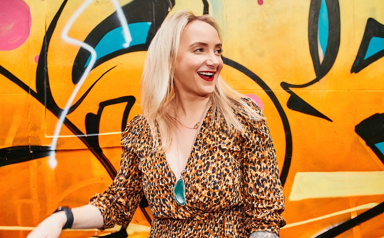 Alicia is standing in front of a bright yellow graffiti wall with flashes of black, blue, and pink. She’s got straight blonde hair past her shoulders, ruby red lips, and is wearing a leopard print long-sleeve v-neck wrap dress. She’s turned towards her left with a wide and cheeky smile. 