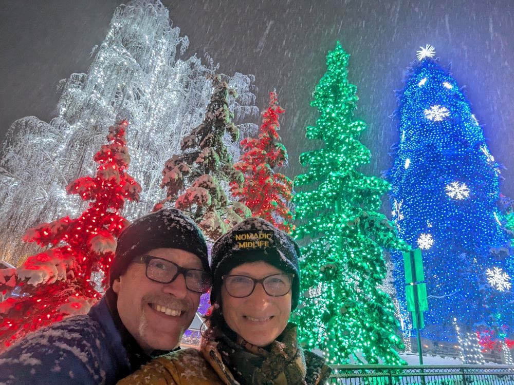 Andy and Sherry in winter coats and hats, posing in front of several large, brightly decorated trees with heavy snow falling against a dark sky