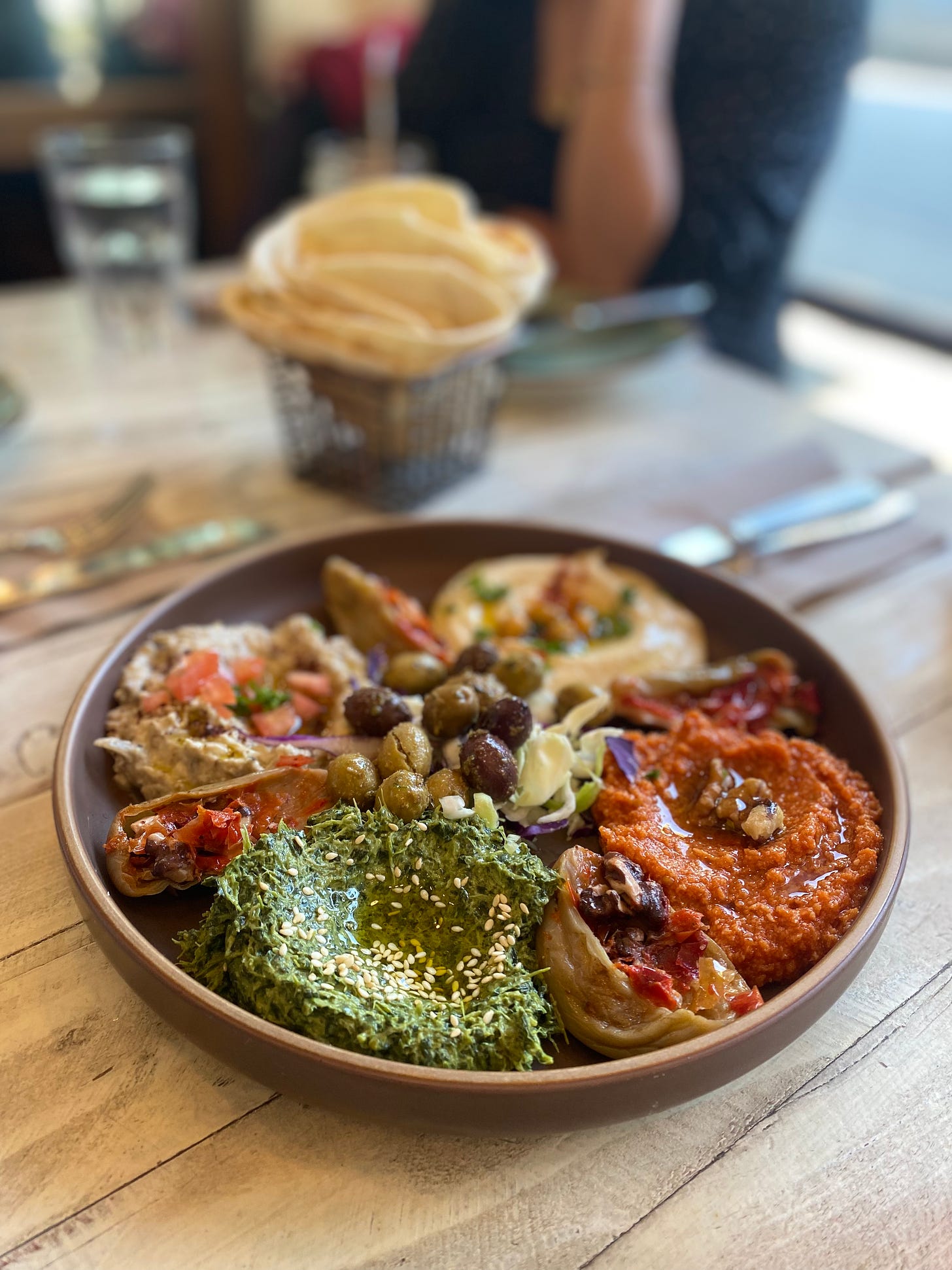 A stoneware plate of neatly arranged Lebanese dips in various colours, with cabbage and olives in the centre.In the background is a wire basket of flatbread.