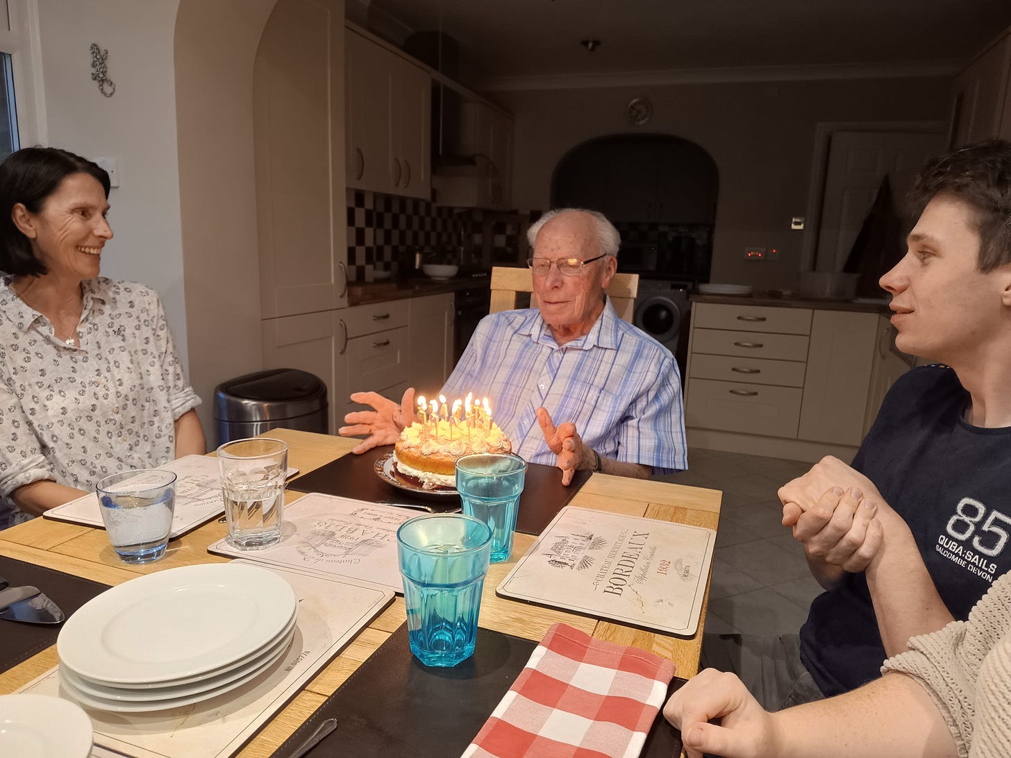 Old man looks surprised at birthday cake with lit candles, surrounded by smiling members of family