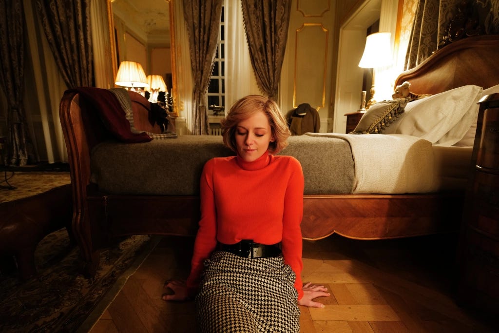 A white woman in a red sweater and checkered skirt sits on the floor by a bed. The woman is blonde and sitting down.