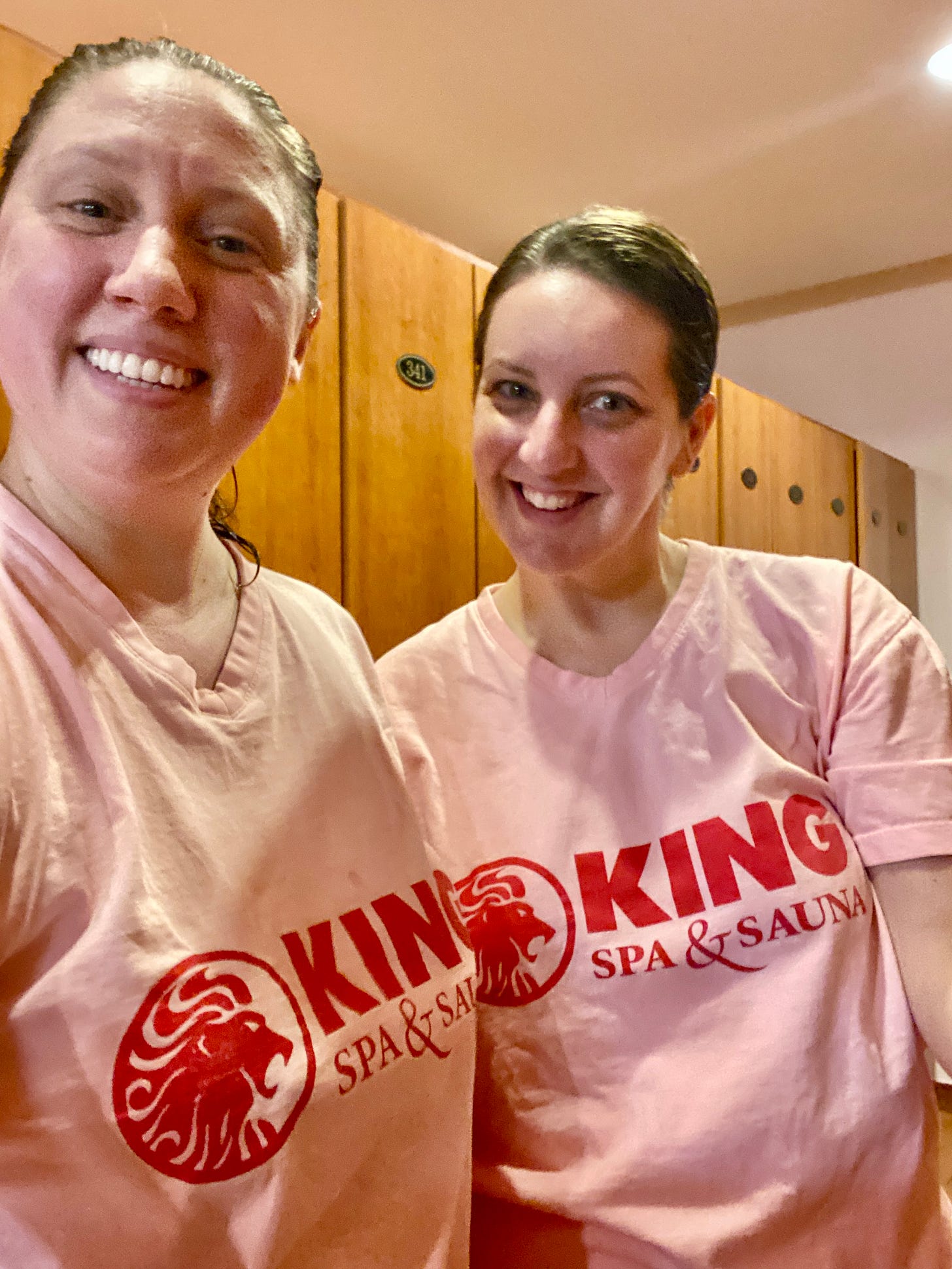 A selfie of two middle-aged whtie women in a locker room (me and a friend). We are wearing pink, v-neck t-shirts that say King Spa & Sauna on the front in red, with the image of a lion's head also in red. We are smiling and our hair is wet and pulled back