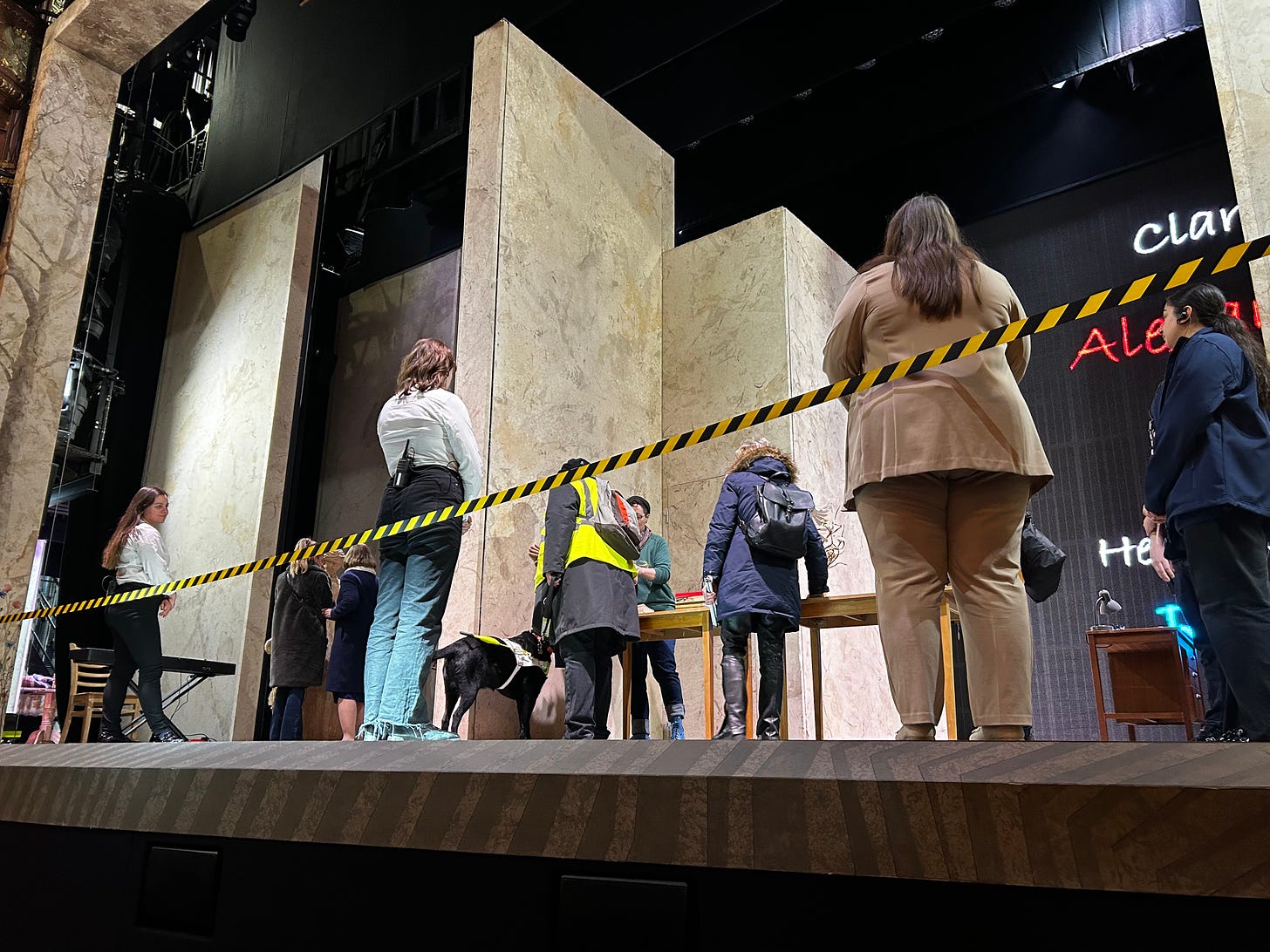 Stage with many people, only backs are visible, man with a guide dog looks at something on a table