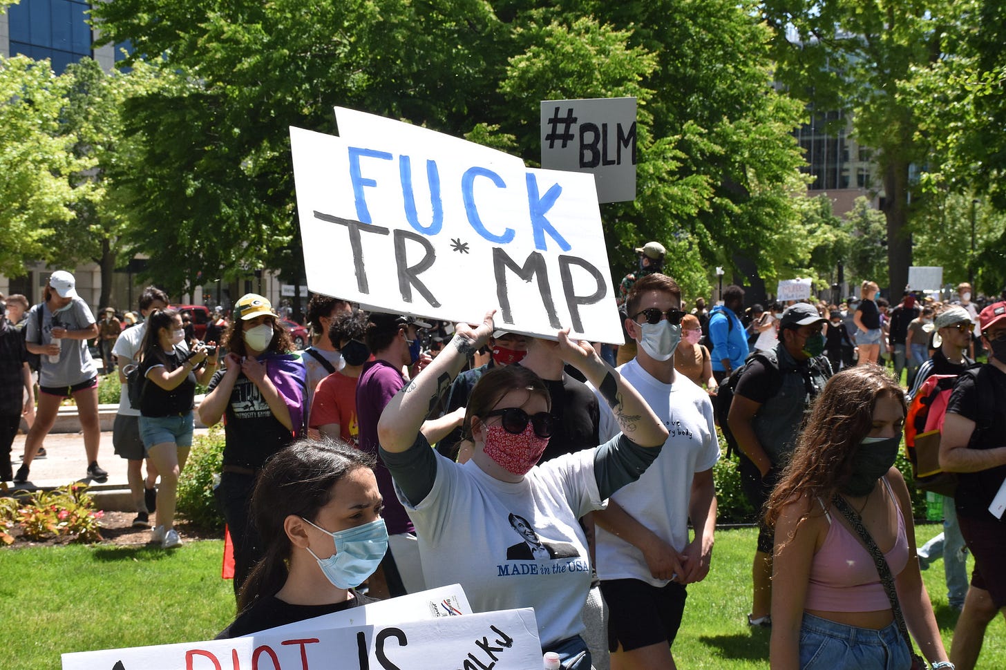 A group of protesters walks through a grassy area, all wearing masks. In the foreground, one person holds a sign that reads "FUCK TR*MP"