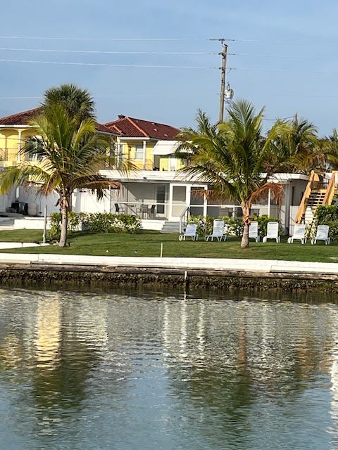 A low slung white house with a screened patio and water in front, framed by palm trees