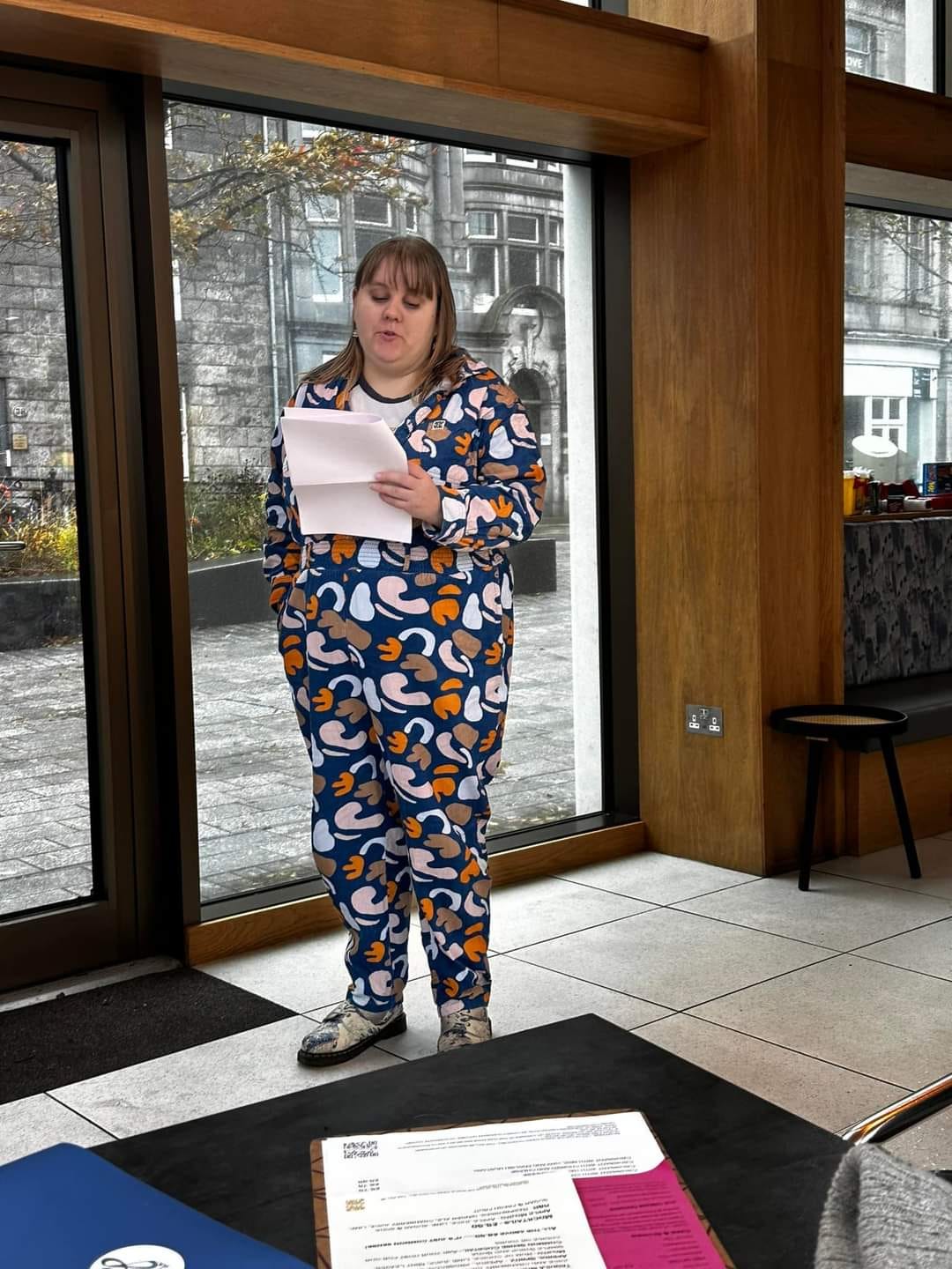 Hannah, 35, is a 5'5.5" white female with blonde hair. She is wearing a blue and autumnally patterned corduroy Lucy and Yak jumpsuit, a Tom Petty t-shirt and a pair of Doc Martens boots with a Great Wave of Kanagawa design on them. She is standing inside a cafe on a dreich Aberdeen day reading poems from a sheet. 