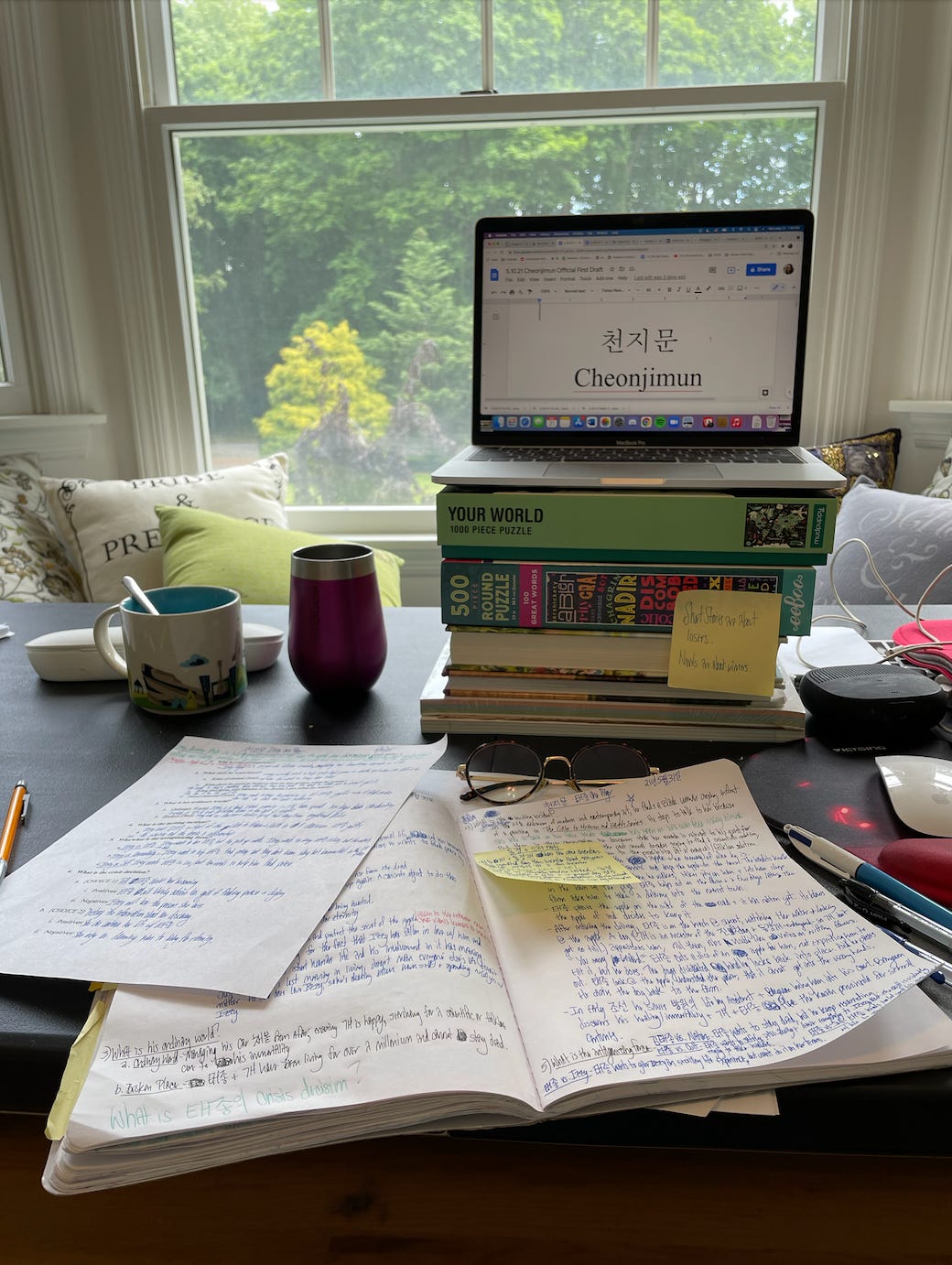 A photo of Kat’s opened notebooks with her messy handwriting and her laptop opened to her novel’s Word doc. Her novel’s title, CheonJiMun is visible on the screen.