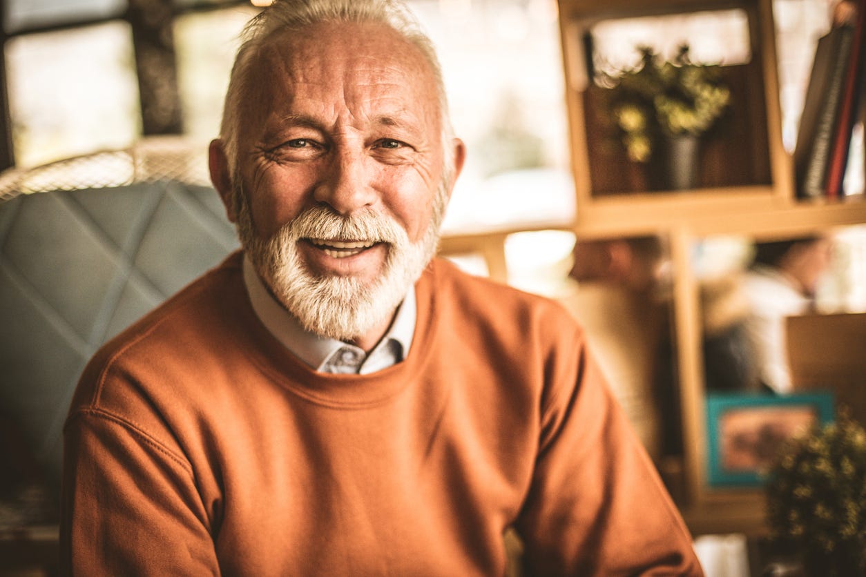 Portrait of smiling white man with white hair, mustache and beard in gray shirt and orange pull-over sweater. 
