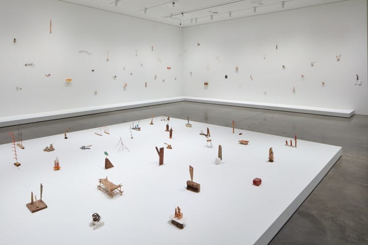 An array of small objects is set up on a low, white platform on a gallery floor. Similar objects are mounted on the wall. The objects are made of wood, string, twigs, and bits of trash, and are barely held together.