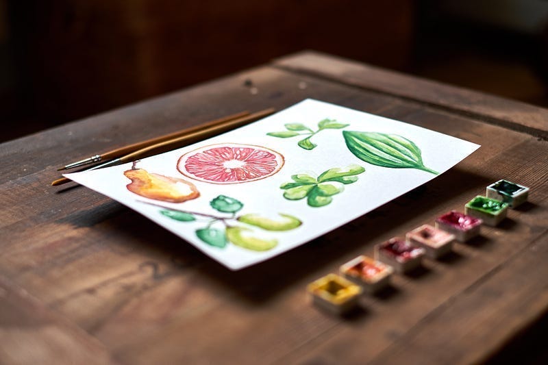water color paints, paintbrushes, and a painting of fruit sitting on a wooden table