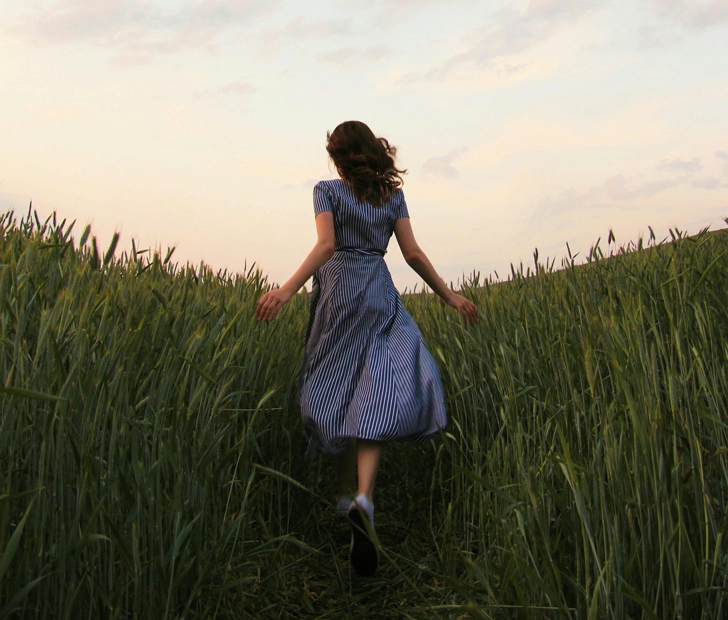 Woman in striped dress running into a field of wheat under a cloudy sky.