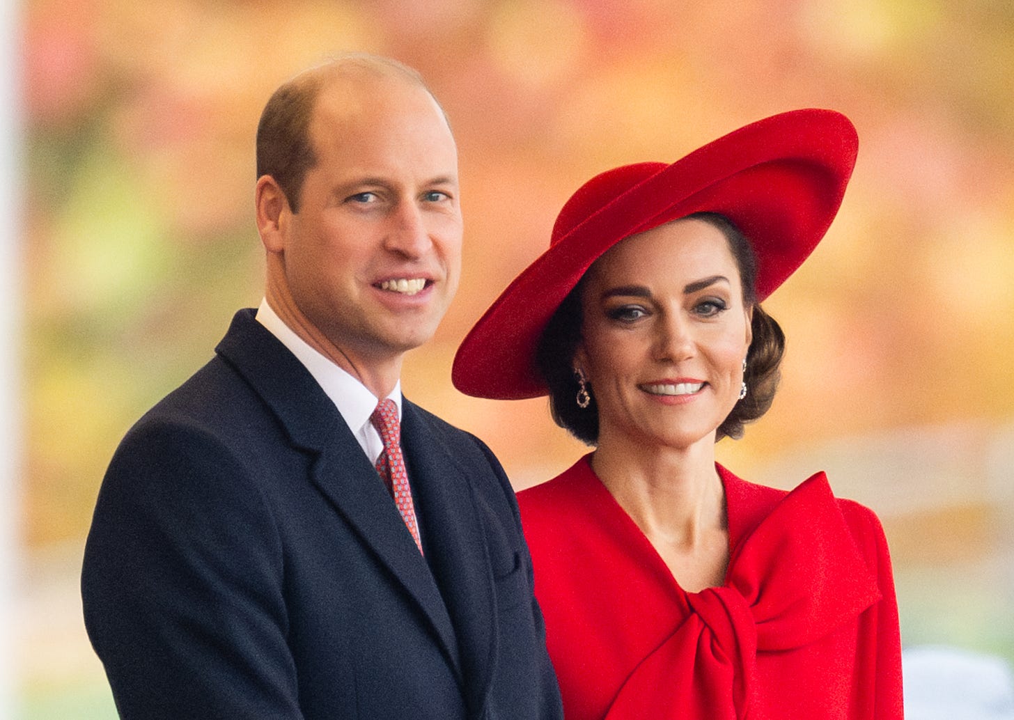prince william on the left smiling in a suit and princess kate in a red dress smiling on the right