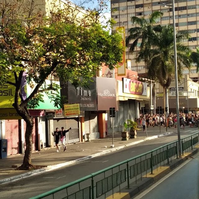 Foto de trecho da Avenida Goiás no ponto em que cruza com a Avenida Araguaia, em Goiânia. A pista está vazia, e as lojas, fechadas. A Avenida Araguaia está repleta de pessoas que andam descendo a avenida, para o lado direito da foto, algumas com bandeiras LGBT. No lado esquerdo, na calçada que acompanha a Avenida Anhannguera, um casal de homems magros vestindo roupas pretas anda lado a lado. Ambos estão abraços parecendo felizes e um deles segura uma caneca. No fundo, se vê prédios, árvores, palmeiras e o céu azul.