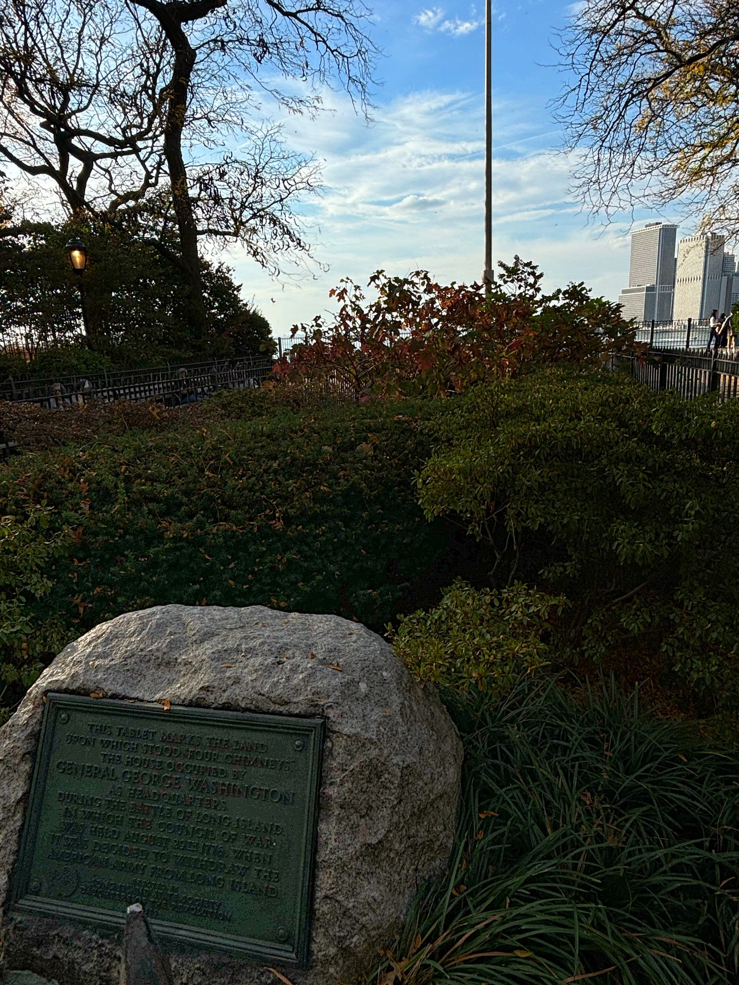 A rock with a metal marker sits in a small fenced in park. Beyond are New Yorkers enjoying the Brooklyn Heights promenade on a warm day. The trees are starting to turn but the sky is blue and the city skyline appears through the trees.