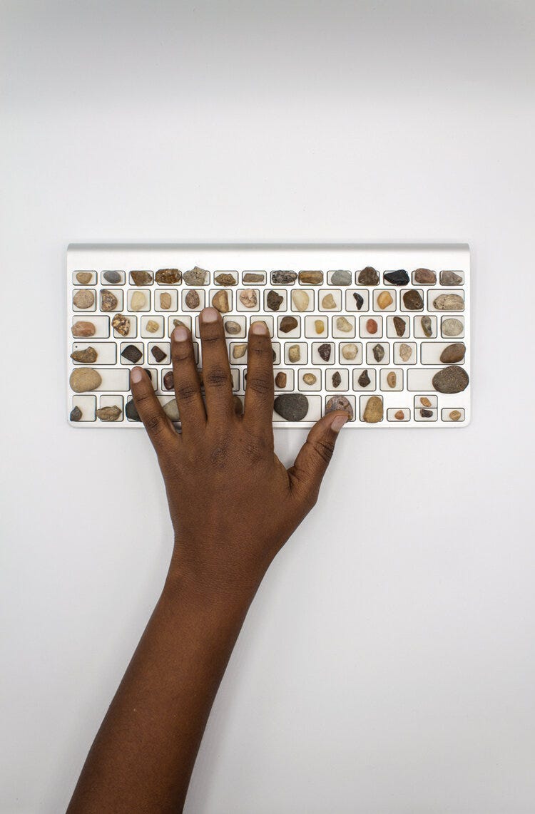 A black persons hand on a computer keyboard with stones on top of the keys.