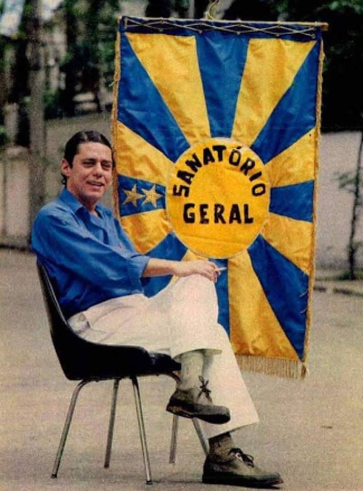 fotografia antiga do chico buarque sentado em uma cadeira ao lado de um estandarte escrito “sanatório geral”. ele está de camisa azul e calças brancas e segura um cigarro. o estandarte é azul e amarelo.