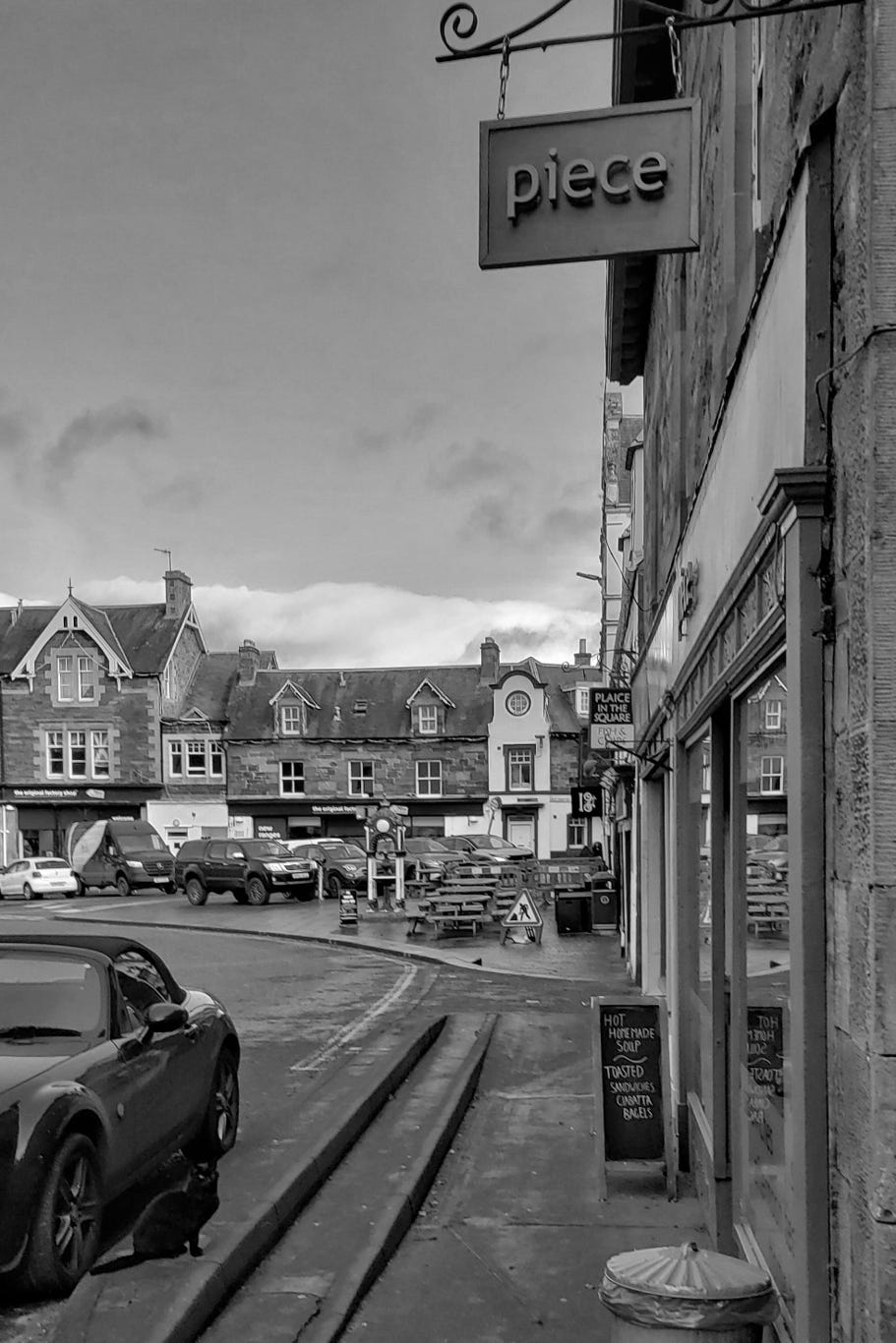 an image of a small Scottish town with a black cat in the bottom left corner staring at the door of a cafe