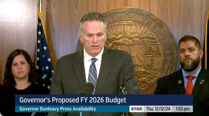 A screenshot of the KTOO Gavel Alaska livestream of Gov. Mike Dunleavy's news conference rolling out the budget. He's standing at the center of the screen flanked by his budget director and revenue commissioner. A carved wooden state seal and flags are in the background.