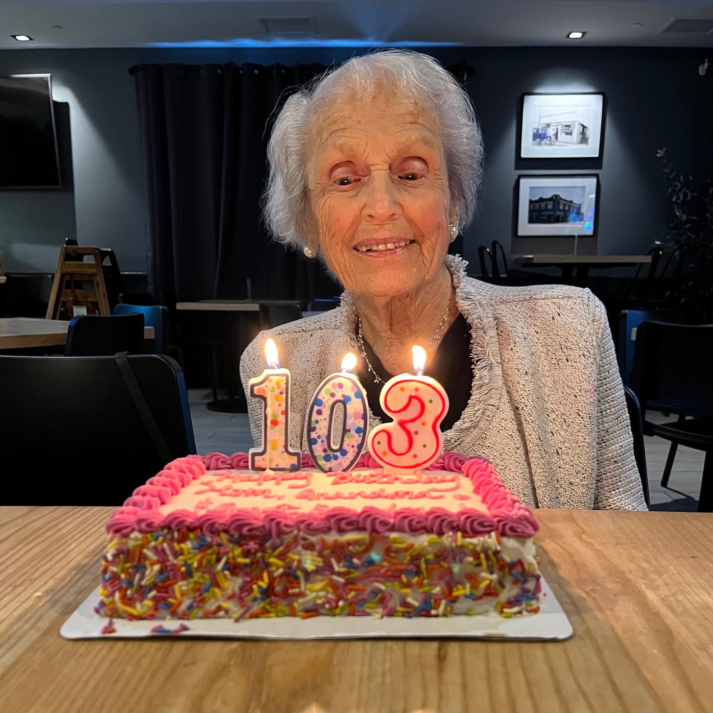 A woman looks down with a smile on a cake decorated with 3 candles "103." Photo by Kevin Ferguson, All Rights Reserved.