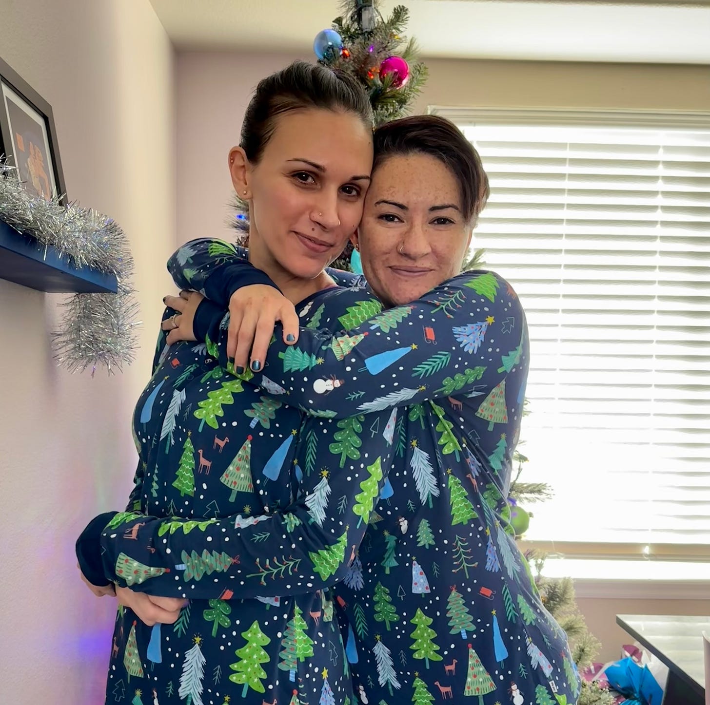 Jessie and Shohreh are in front of the Christmas tree wearing matching blue PJs with a pattern of green Christmas trees on them. Jessie's arms are slung around Shohreh's neck, and they have their heads together