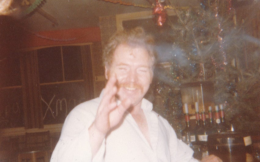 A faded 1980s photo of a man with red hair and red moustache smiling, perhaps a little tipsy, in a pub.