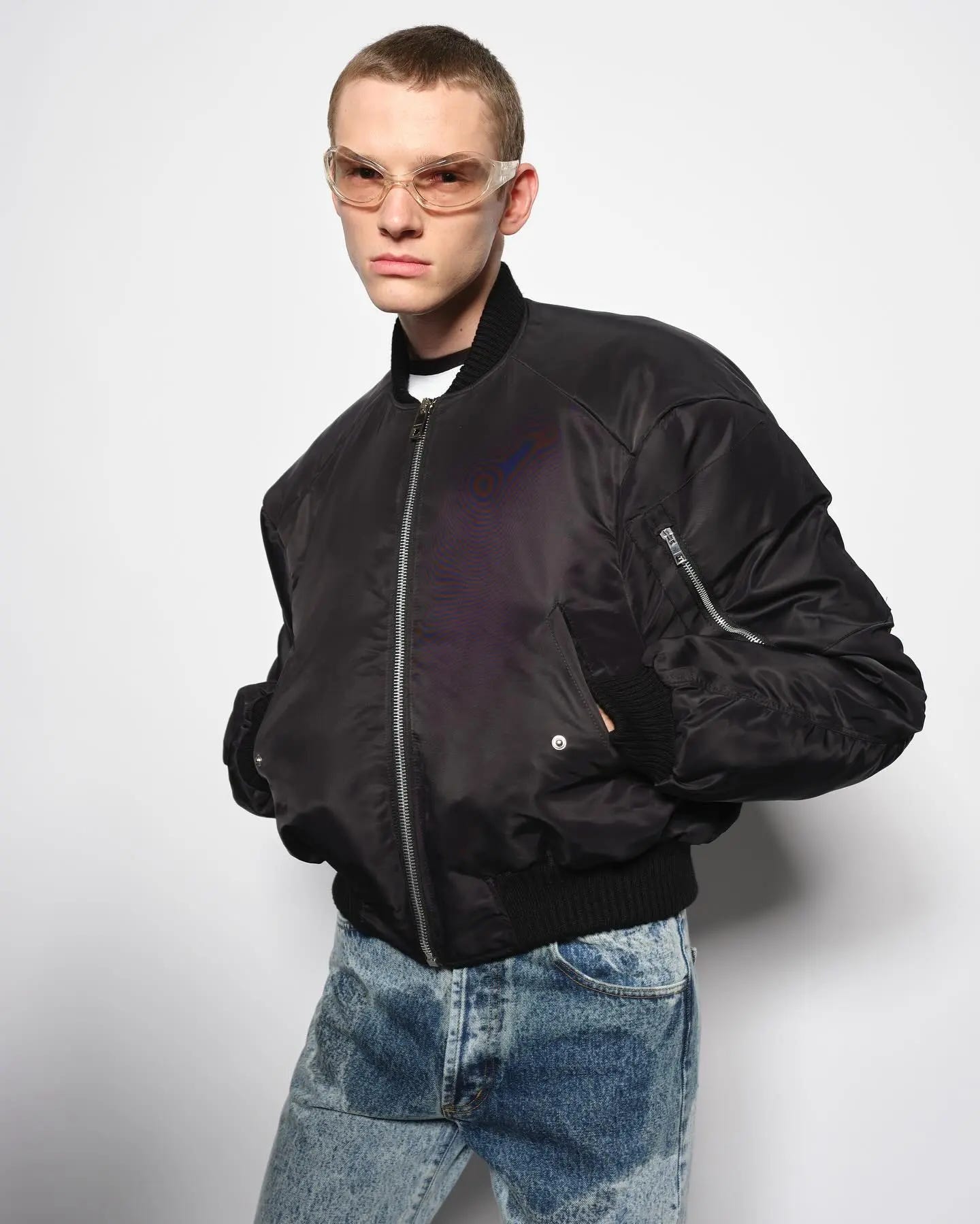 A guy in a black bomber jacket wearing light denim. The crotch of the denim is dark, making it look wet -- nearly all the way up to the pockets. Also he's wearing a pair of glasses that look like safety glasses. Modeling.