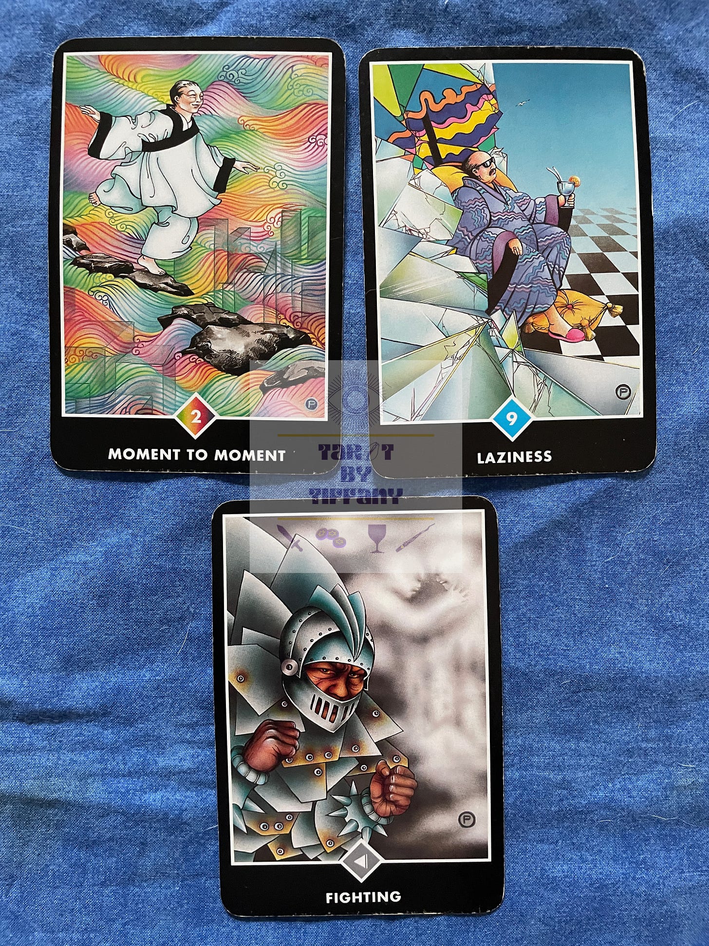 3-card reading using the Osho Zen Tarot. The cards are laid on a cobalt blue cloth background. Top two cards (L-R): 2 of Rainbows - Moment to Moment, 9 of Water - Laziness. Bottom card: Knight of Clouds - Fighting.