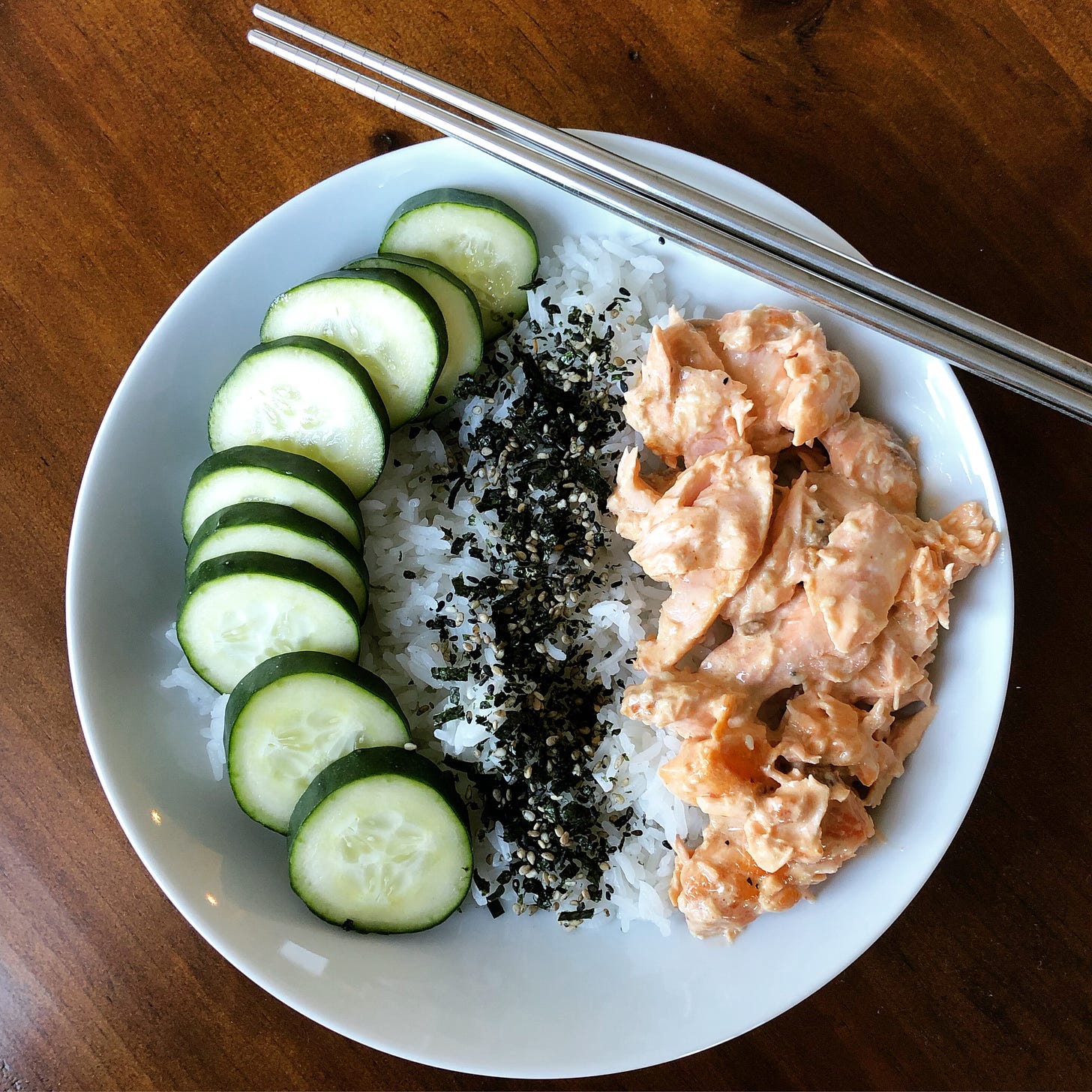 A bowl with rice topped with sliced cucumbers, furikake, and spicy salmon.