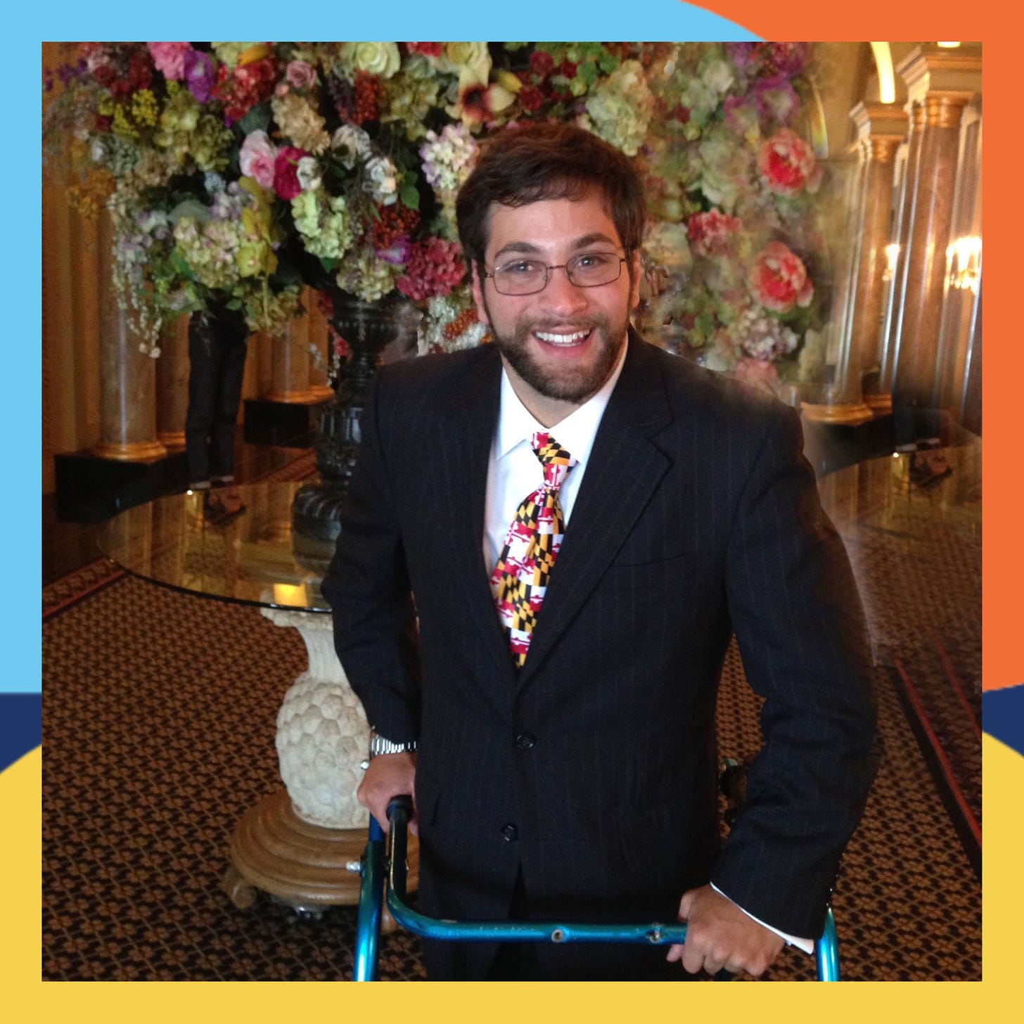 Aaron wears a suit and tie, He has brown hair and a mustaches and goatee. He uses a walker and smiles at the camera.