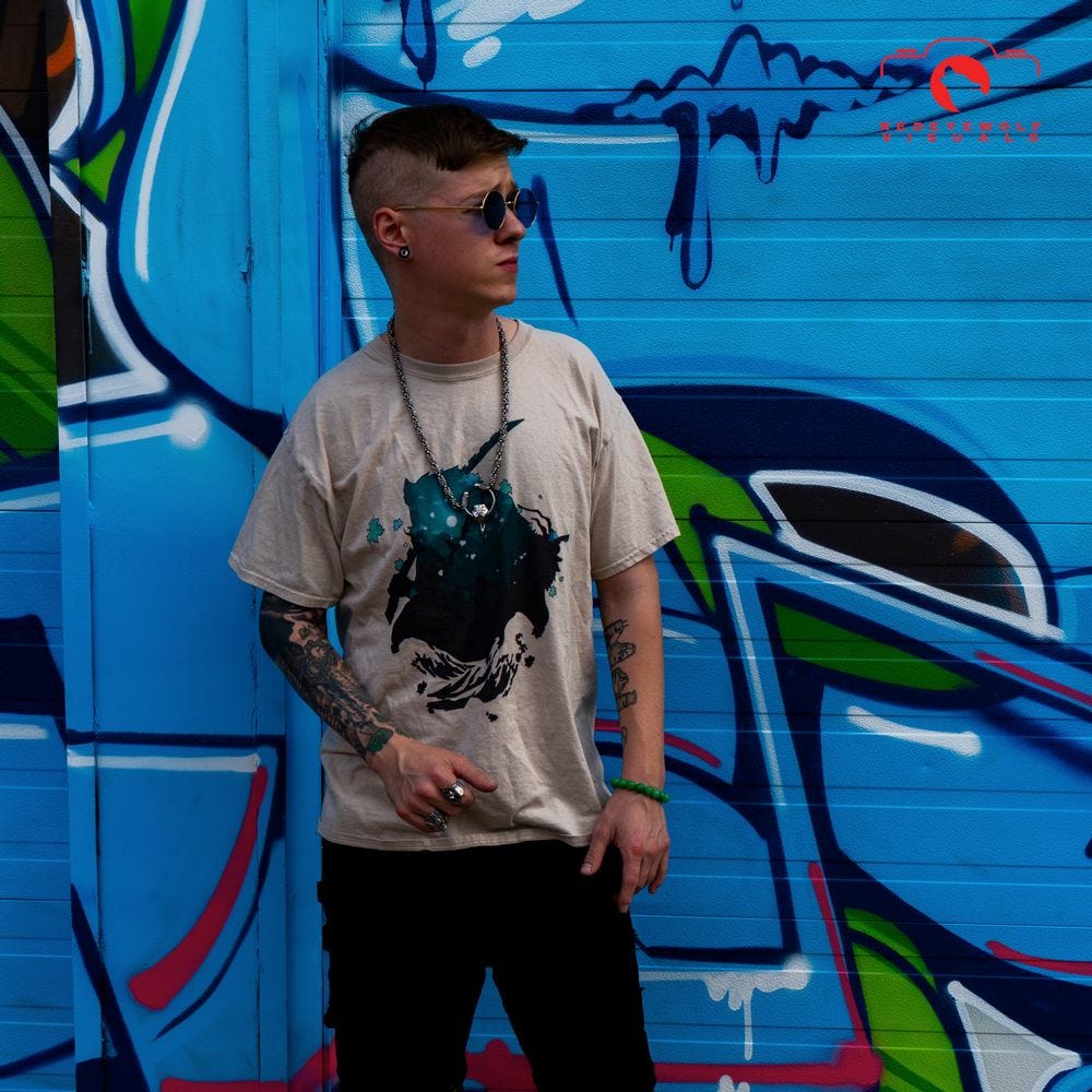 Crowfather, a young white man with arm tattoos, a high & tight haircut, and heavy jewelry, looks to the side, while standing in front of an artfully graffiti'd wall.