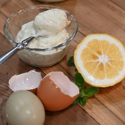 A bowl of mayo with a spoon, cracked eggs and a halved lemon on a counter.