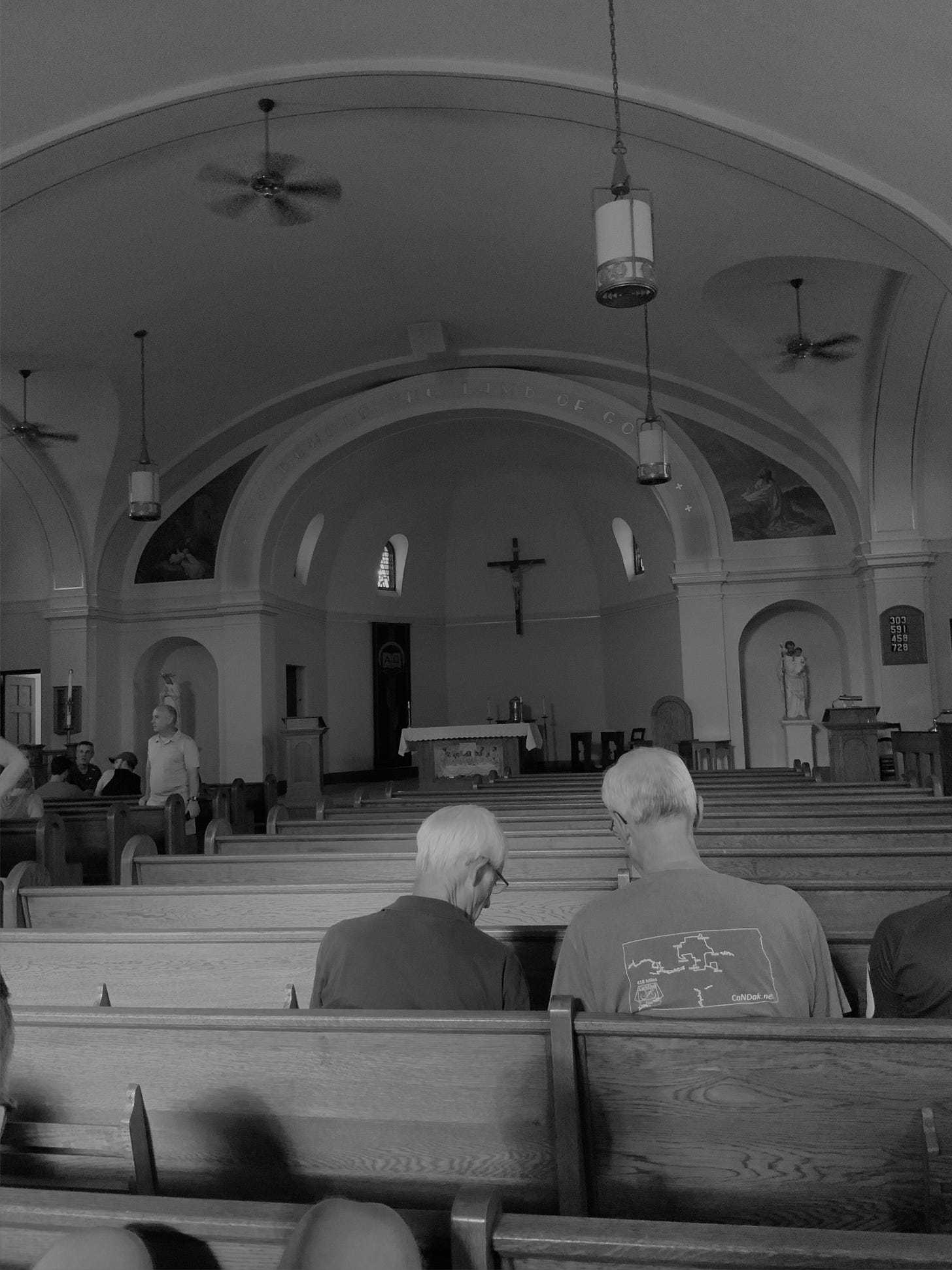 The interior of a Catholic church.  Two older gentlemen are in a pew ahead of me, and several others are seated toward the front of the church, but otherwise the church is empty.  A recessed, domed area is there for the altar and crucifix; occasional statues and paintings decorate the church.  Fans hang from the ceiling.