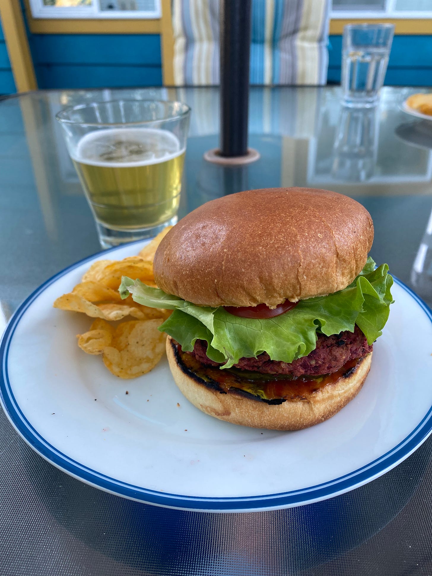 A white plate with a blue rim on a glass patio table; on the plate is a beet burger on a brioche bun with lettuce, tomato, and pickles just visible. Next to the burger is a small pile of spicy pickle chips, and behind the plate is a glass of lager.