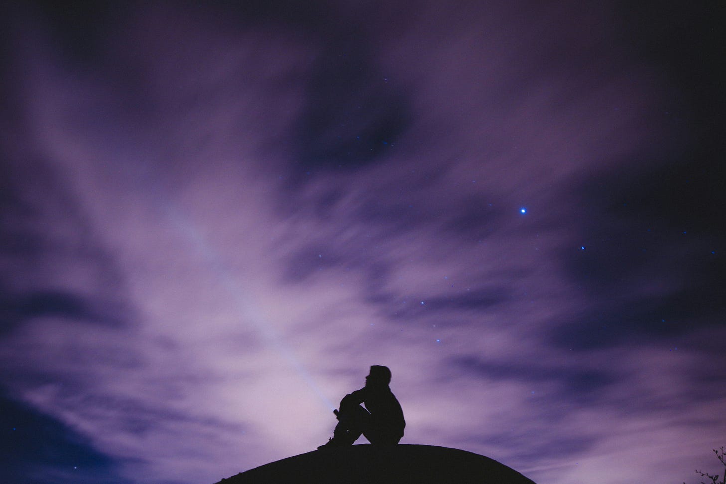 person sits on a hill underneath a violet dark sky with stars