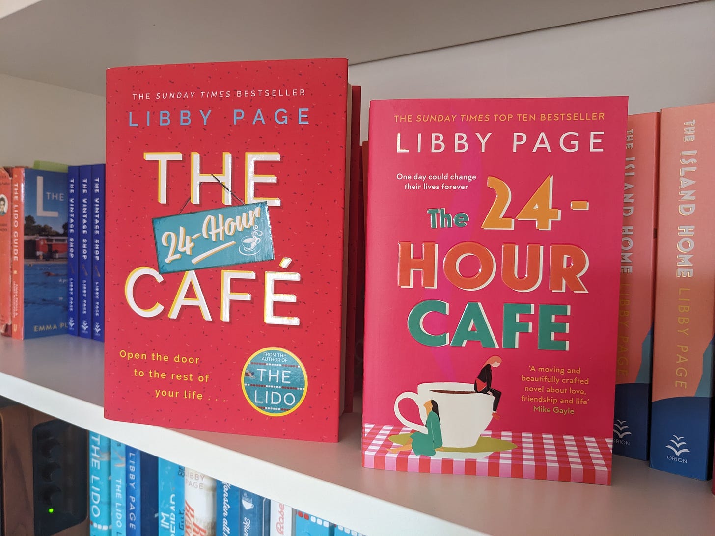 The hardback of The 24-Hour Cafe by Libby Page next to the paperback, on a bookshelf. The hardback is dark red and text based. The paperback is a deep pink and features an image of two women sitting near a teacup on a table with a red and white check tablecloth.