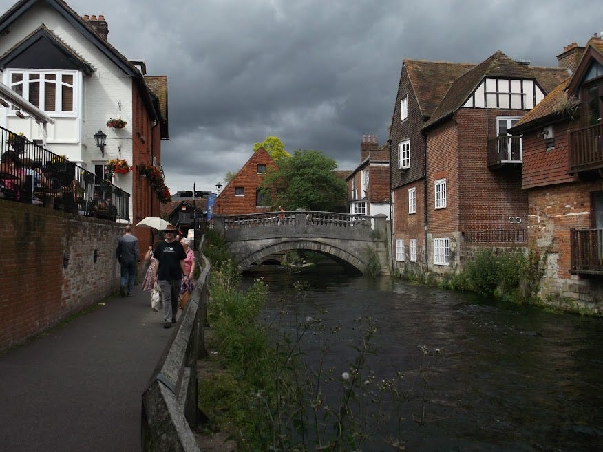 Picture of a river through a city under a stone bridge. A pub is on the side. People are walking alongsied the river on a fenced footpath.