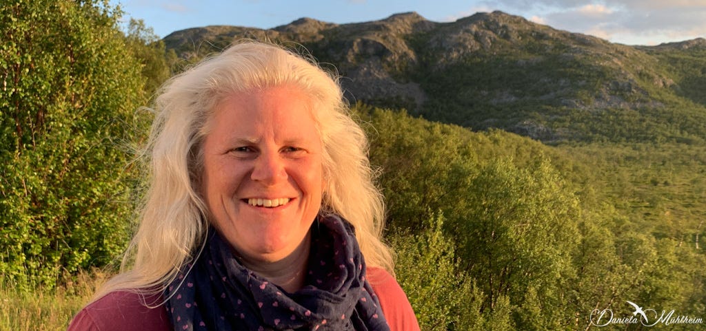 Daniela Mühlheim smiling happily in a portrait shot with norwegian mountains and trees in the background wearing a marine blue scarf and a ruby coloured shirt