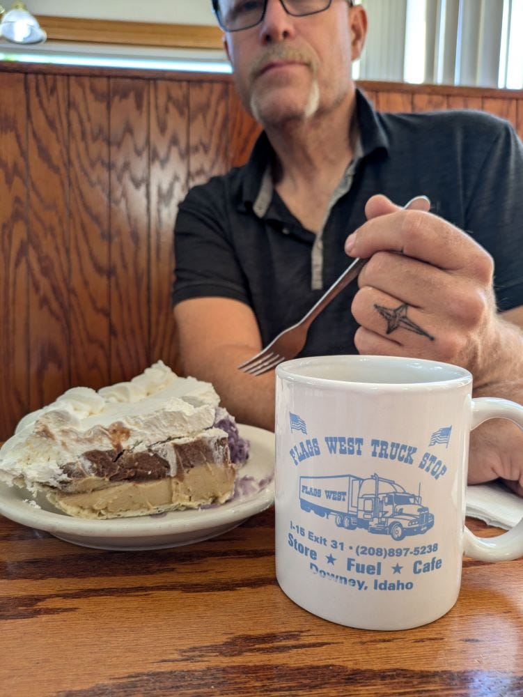 Andy in a restaurant booth, looking on at a slice of pie, with a coffee mug bearing the name of the truck stop in Idaho