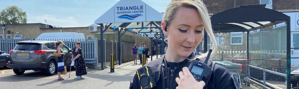 Female officer using radio outside Triangle shopping centre entrance.