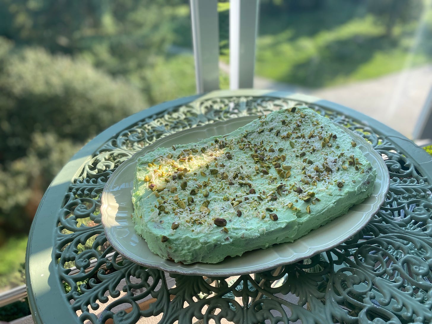 A café table with a large platter on it holding the Watergate cake, garnished with pistachios and shredded coconut. All of it is a similar shade of mint green.