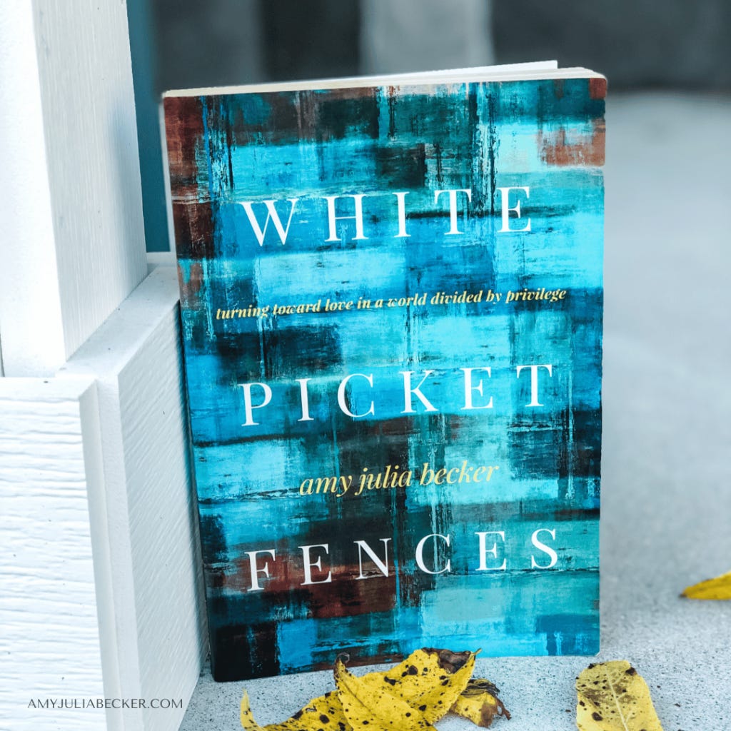 The image shows a book titled White Picket Fences by Amy Julia Becker. The cover features an abstract design in various shades of blue with white text. The subtitle reads, "Turning toward love in a world divided by privilege." There are yellow autumn leaves scattered near the book, and the background has a soft, blurred gray tone. The author's website, "amyjuliabecker.com," is printed in the bottom left corner.